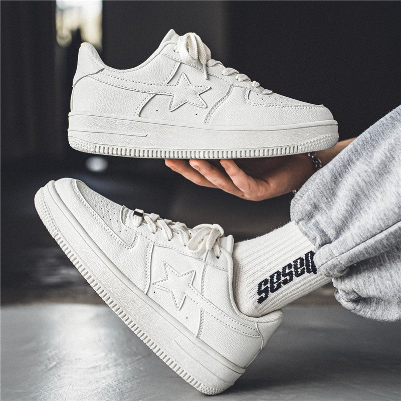 A pair of white low-top sneakers with embossed star details on the sides, shown with one shoe held in a hand and the other resting on a surface. The person is wearing a gray sweatpants leg and a white sock with black text.