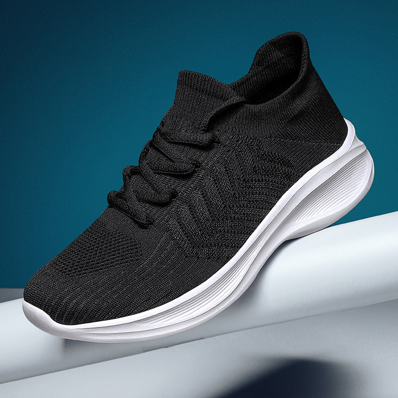A black knit slip-on sneaker with a thick white sole, resting on a white cylindrical platform against a dark teal-blue background.
