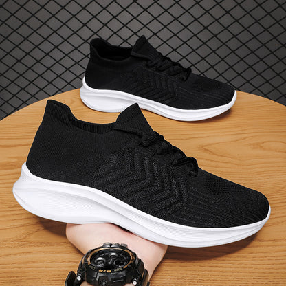 A pair of black knit slip-on sneakers with thick white soles, displayed on a wooden table with a hand holding one shoe and a watch visible in the foreground.