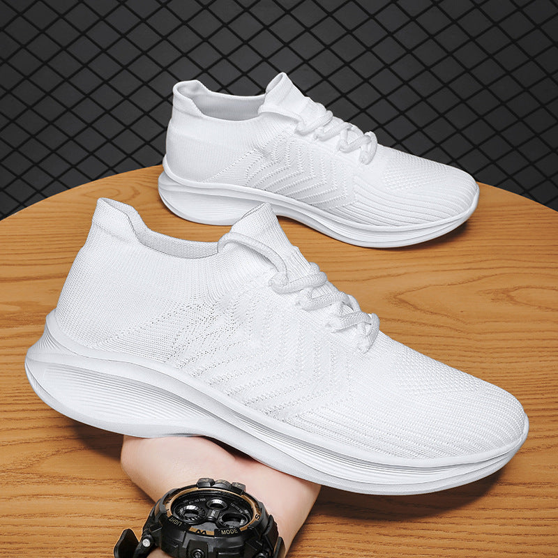 A pair of white knit athletic sneakers with thick white soles, displayed on a wooden table with a person's hand and a black watch visible in the foreground. A dark textured background is behind the table.