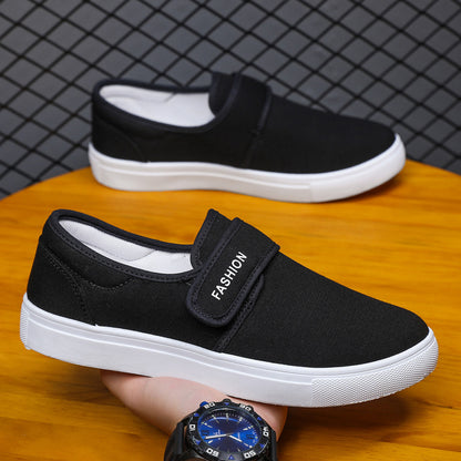 Black slip-on shoes with white soles on a wooden surface, held by a hand wearing a watch.