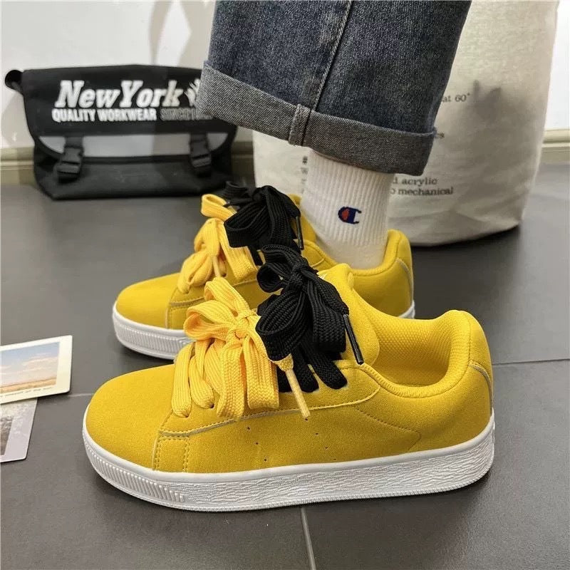 Yellow sneakers with black laces worn by a person, with a gray surface and bags in the background.